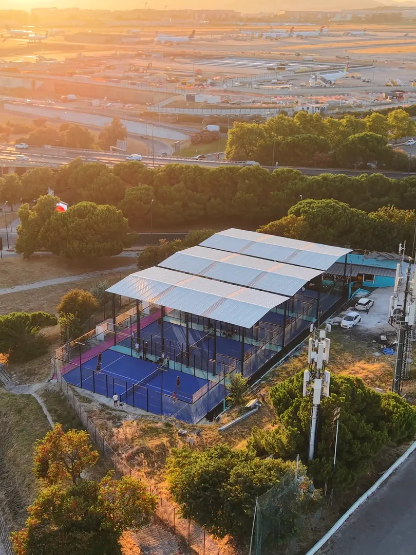 O Padel do Relógio visto de drone ao pôr do sol: os campos azuis sob a cobertura branca do clube, com as pistas do aeroporto de Lisboa e a serra douradas pelo sol ao fundo.